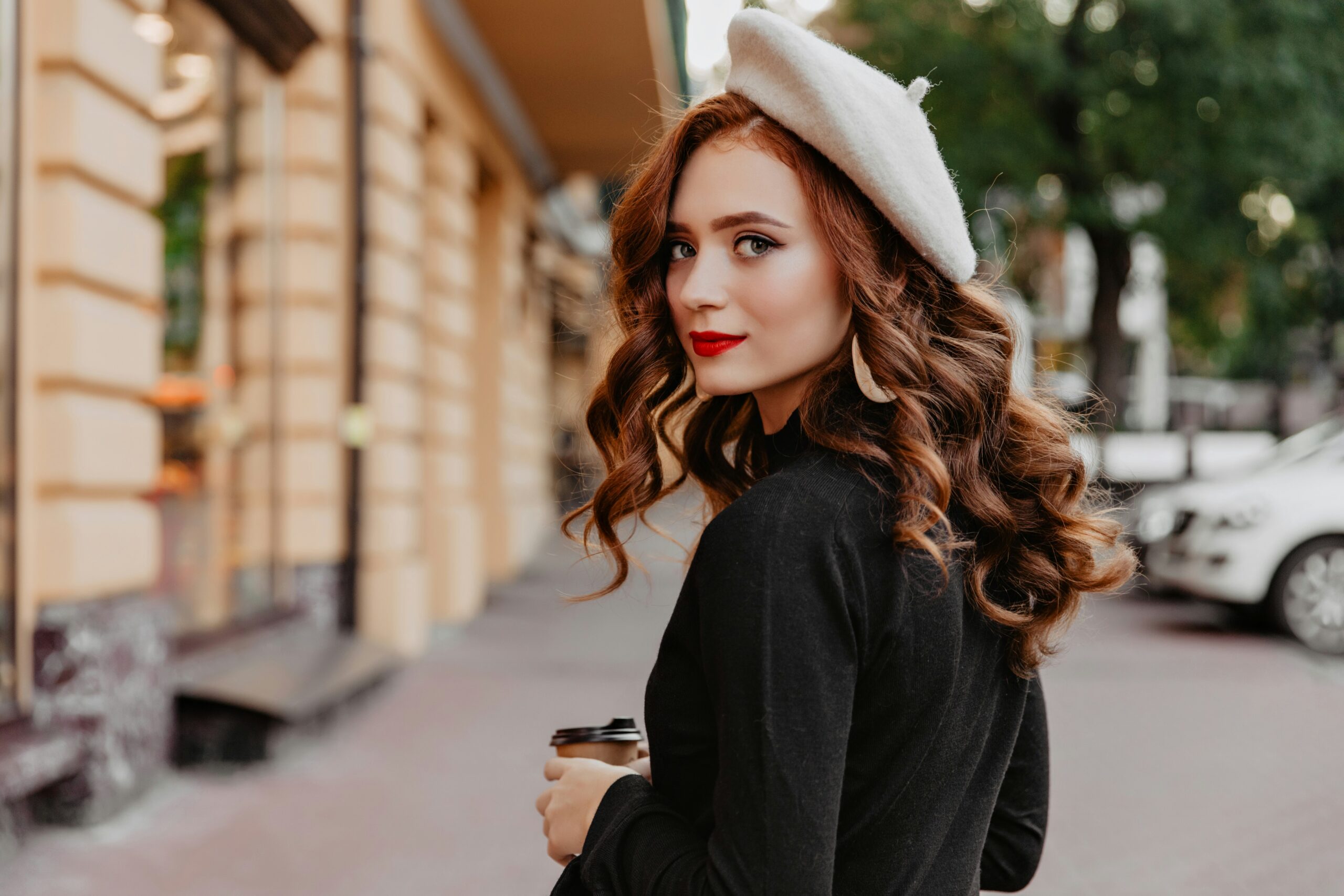 Woman with long, wavy auburn hair and warm copper tones wearing a white beret outdoors.